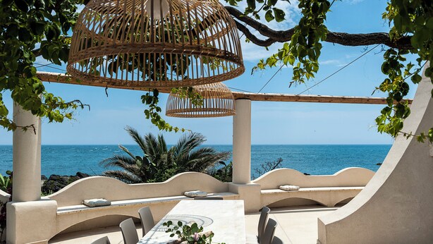 An outdoor dining area with a view of the sea, featuring a table with chairs, a wicker hanging light, and a bench with cushions. - Marina dei Greci (Giardini Naxos)