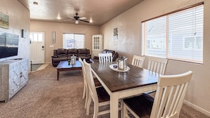A spacious living room with a dark wooden dining table, white chairs, a leather sofa, and a ceiling fan.