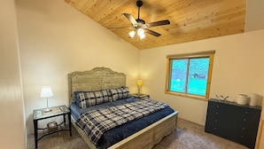 Een slaapkamer met een houten plafond, een bed met geruit beddengoed, een nachtkastje met een lamp, een dressoir en een raam met uitzicht op groen.