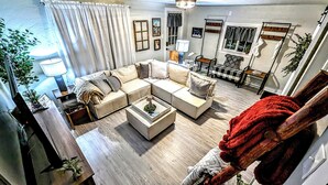 A spacious living room with a large L-shaped sofa, a coffee table, and a red throw blanket. There is a wooden floor, a lamp, and a window with curtains.