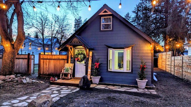 A small, dark-colored house with a front porch, a wooden chair, and a potted plant.