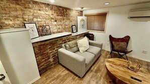 A compact living space with a brick wall, a grey sofa, a wooden dining table, and a red chair. - Downtown SLC 1BR · Marmalade · Walk to Capitol (Salt Lake City)
