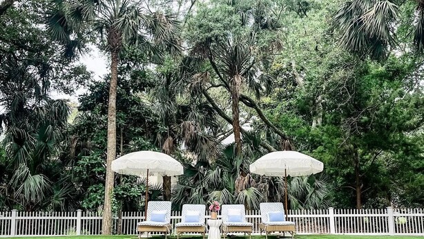 Un área de piscina con sillones de descanso y sombrillas, rodeada de árboles.