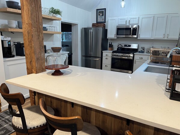 A kitchen with a central island, wooden chairs, white cabinets, stainless steel appliances, and a hanging light fixture.