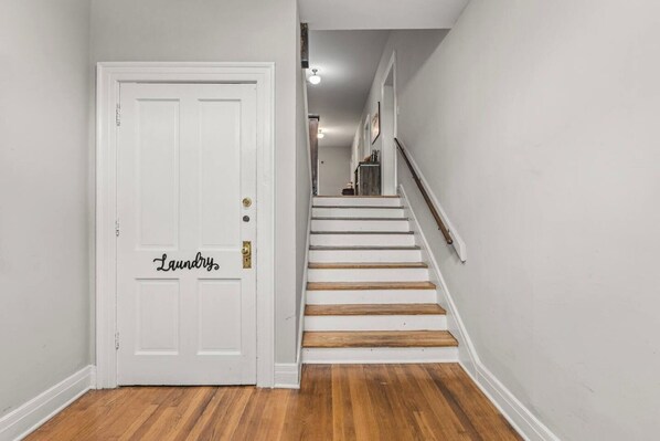 Un couloir avec une porte blanche portant l’inscription « Buanderie » et un escalier en bois menant à l’étage supérieur.