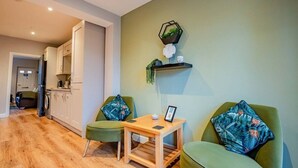 Ein modernes Wohnzimmer mit Couchtisch aus Holz, zwei grünen Sesseln, einem kleinen Beistelltisch und einer Wandregal mit Dekoration.