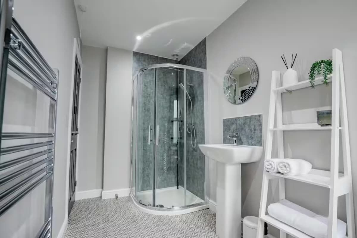 Une salle de bain moderne avec une douche à parois de verre, un lavabo sur colonne blanc et une étagère blanche où sont rangées des serviettes.