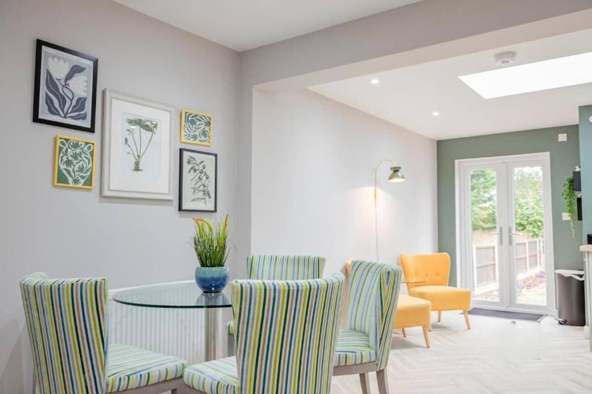 A modern living room with a round glass table, striped chairs, and a yellow armchair. There are framed botanical prints on the wall and a floor lamp.