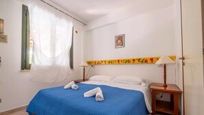 Un dormitorio con cama, mesita de noche, lámpara, ventana con cortinas y un cuadro colgado en la pared.