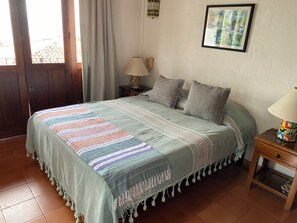 A bedroom with a bed, pillows, a nightstand, a lamp, a framed picture on the wall, and a door with curtains.