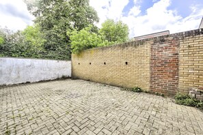 Un patio empedrado con un muro de ladrillo, una pared blanca y un árbol al fondo.
