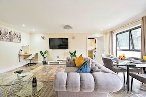 A modern living room with a sofa, a flat-screen TV, a dining area with a table set for two, and a large window. - Bright flat, ideal for contractors, sleeps 5! (Greater London)