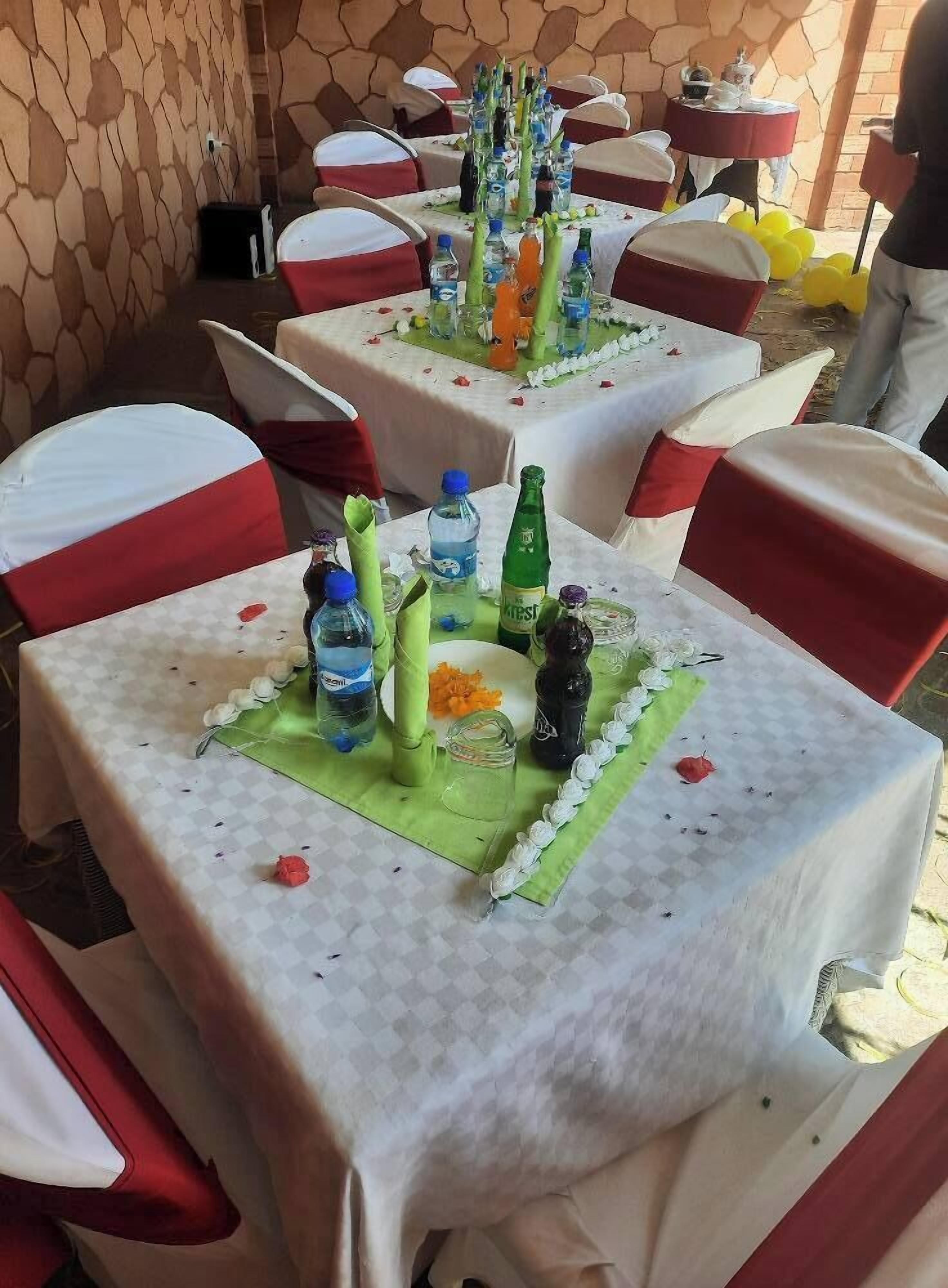 Ein Außenbereich mit Essbereich, Tische sind mit weißen Tischdecken und roten Sitzkissen ausgestattet. Auf jedem Tisch liegen grüne Platzsets, Wasserflaschen und eine kleine Dekoration.