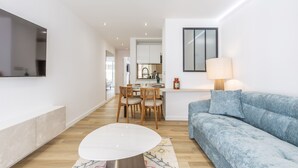 Ein modernes Wohnzimmer mit einer Couch, einem Esstisch mit Stühlen, einem an der Wand befestigten Fernseher und einer Küchenzeile im Hintergrund.