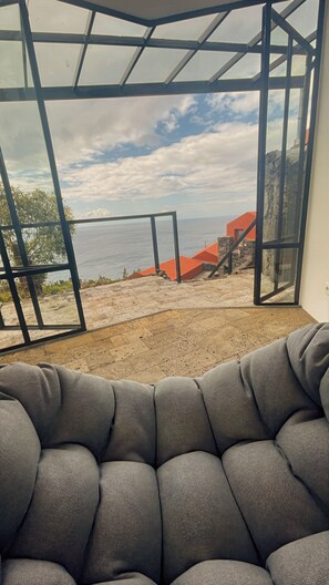A room with a view of a rocky coastline, a glass door leading outside, and a grey upholstered sofa.