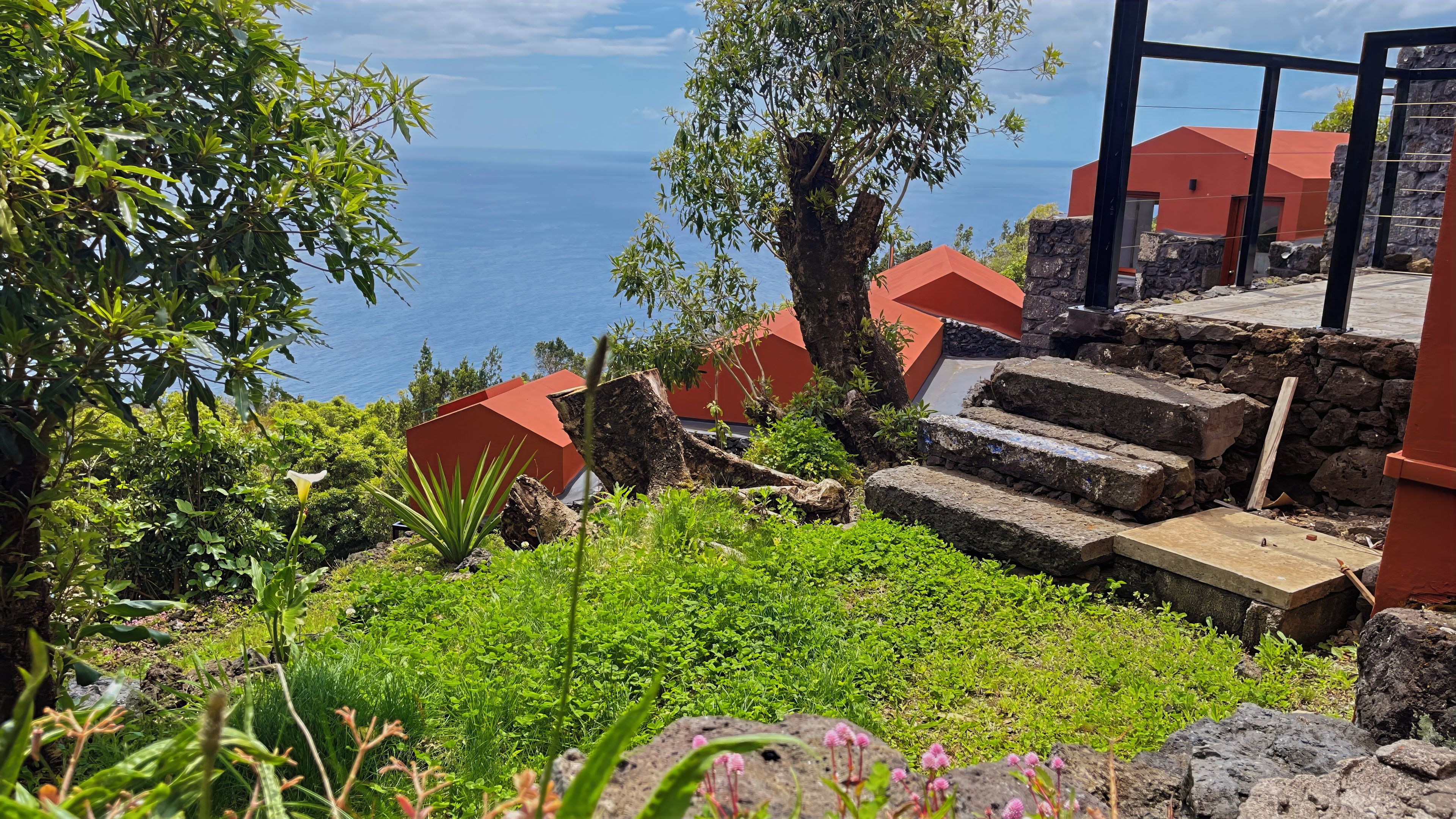 A coastal property with a stone staircase leading to a red-roofed building, surrounded by lush greenery and overlooking the sea.