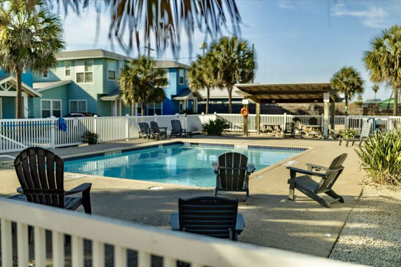 A swimming pool with lounge chairs and a covered area, surrounded by palm trees and buildings.