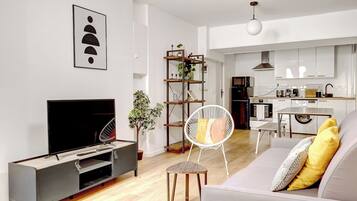 Een moderne woonkamer met een flatscreen televisie, een bank met kussens, een klein houten tafeltje en een witte stoel.