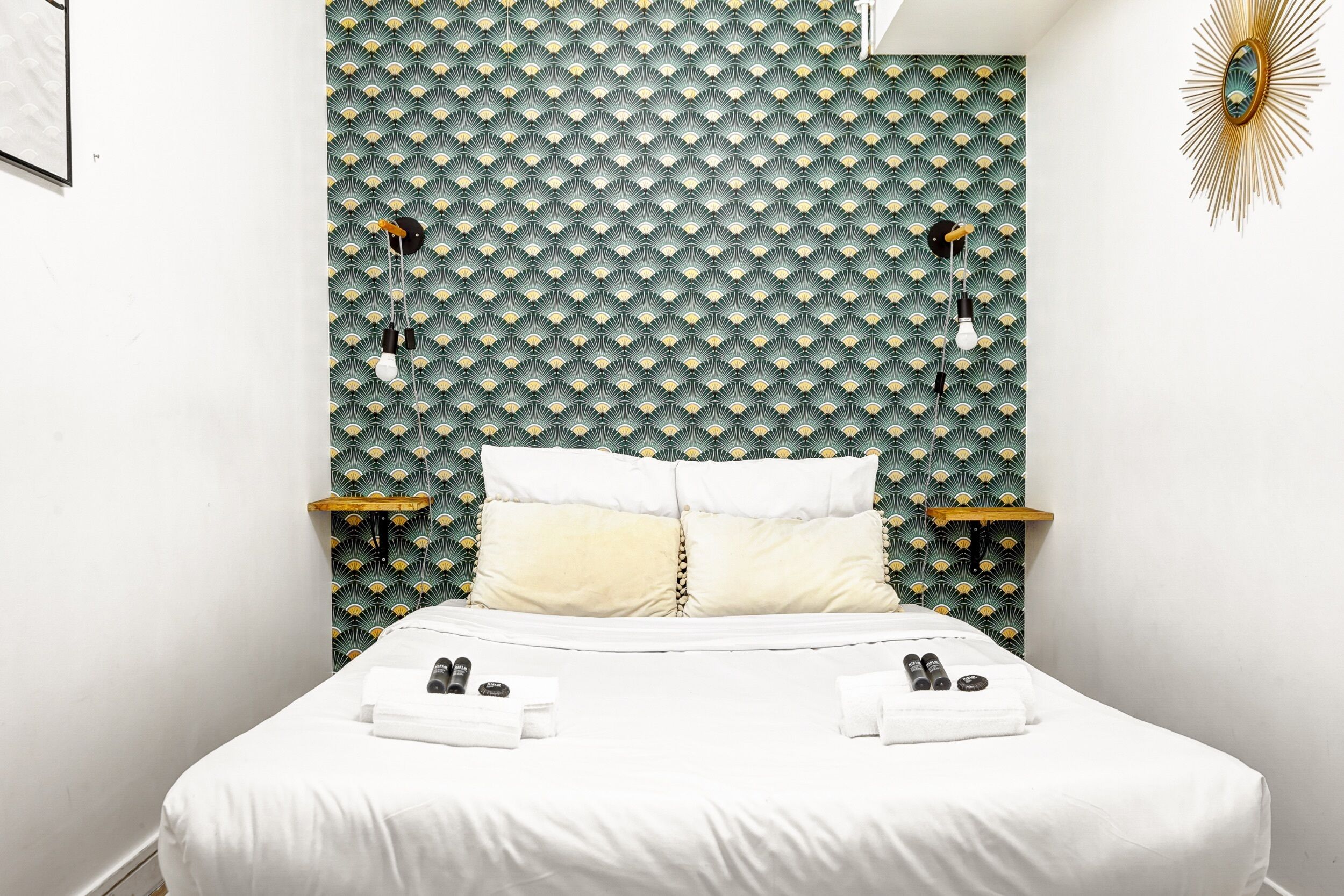 Un lit bien fait, avec du linge de lit blanc et deux oreillers, une table de chevet avec une lampe, et un mur orné d’un motif géométrique.
