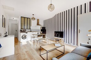 A modern kitchen with a washing machine, a dining area with benches, a coffee table, and a TV in a living room with striped wallpaper.
