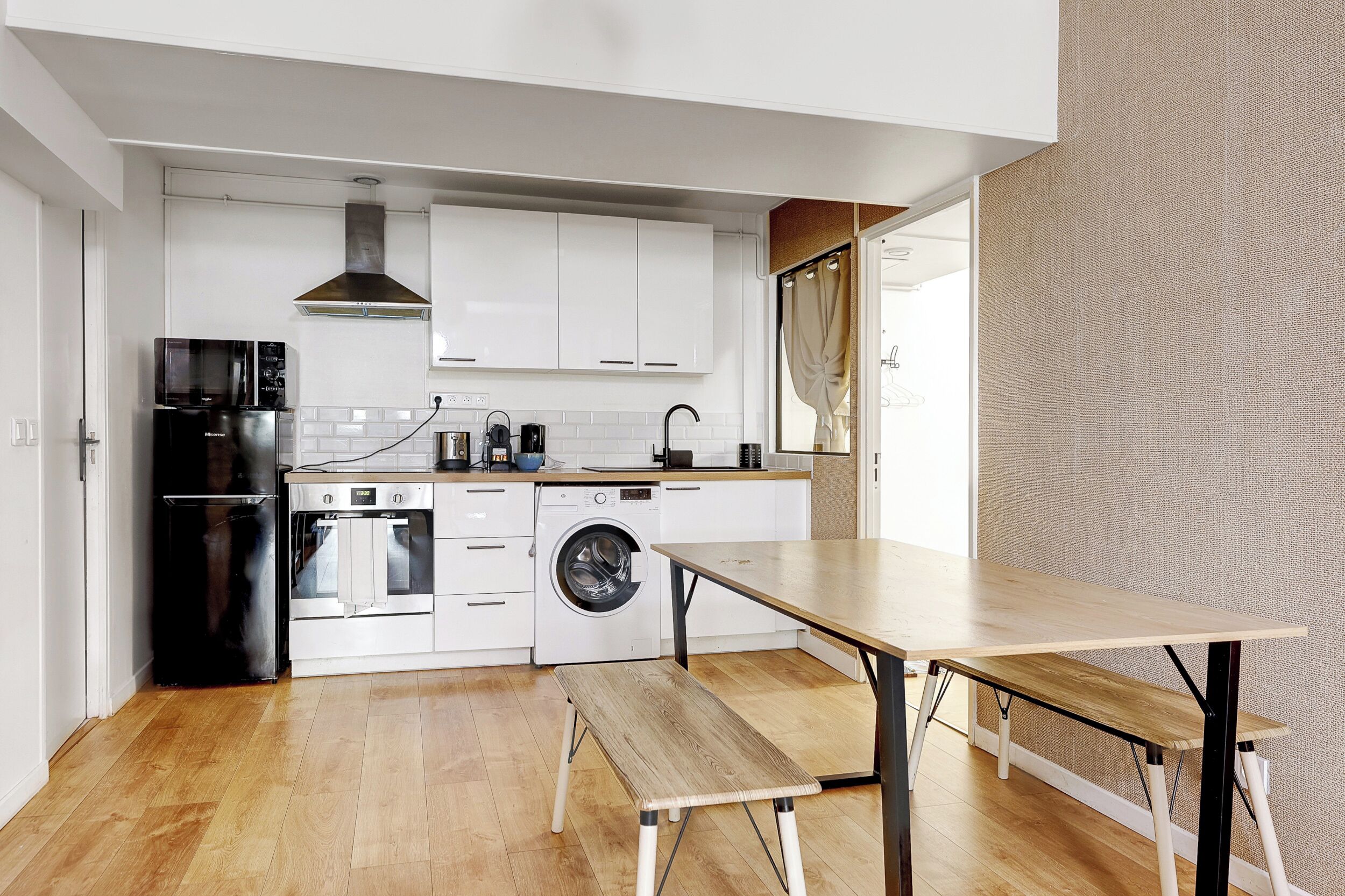 A modern kitchen with white cabinets, a black refrigerator, a stove, a washing machine, and a wooden bench.