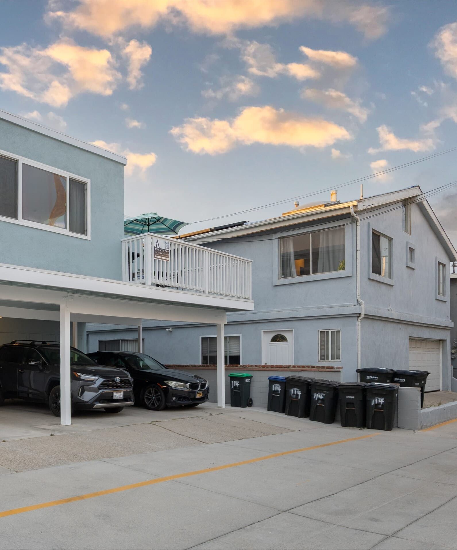 A two-story residential building with a balcony, a garage with two cars, and several trash bins.