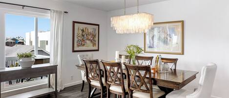 A dining area with a wooden table, white upholstered chairs, a hanging light fixture, and framed artwork on the walls.