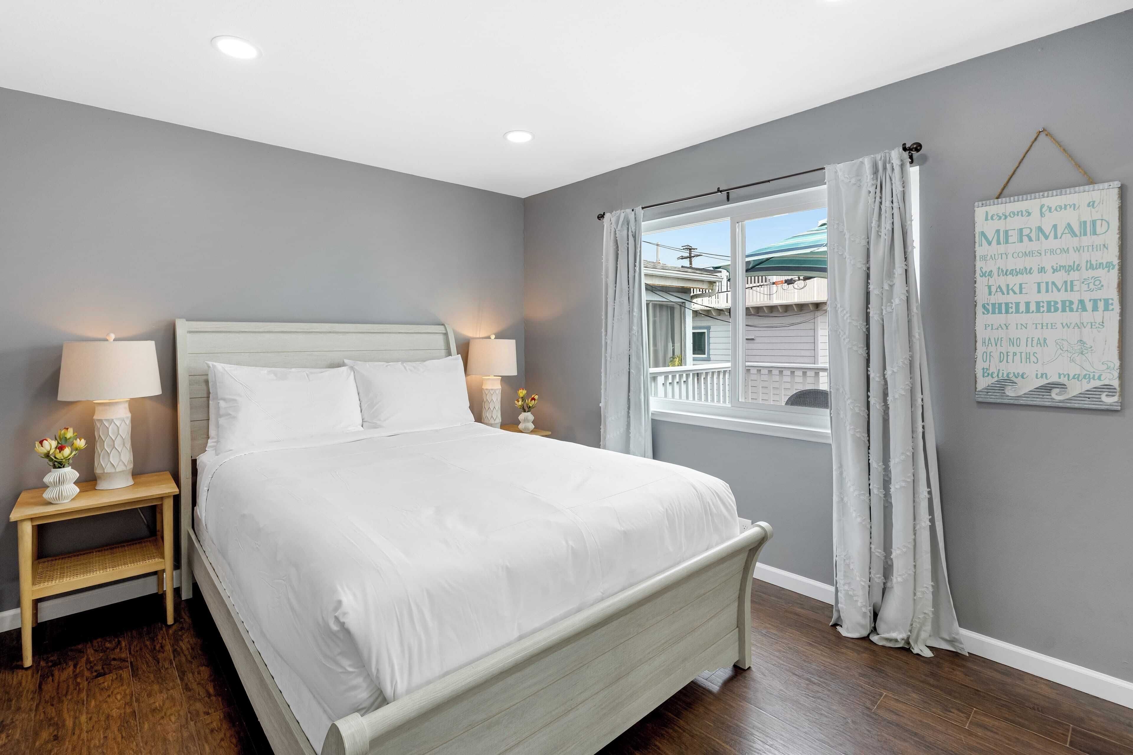 A bedroom with a white bed, two bedside tables with lamps, a window with curtains, and a wall with a motivational quote.