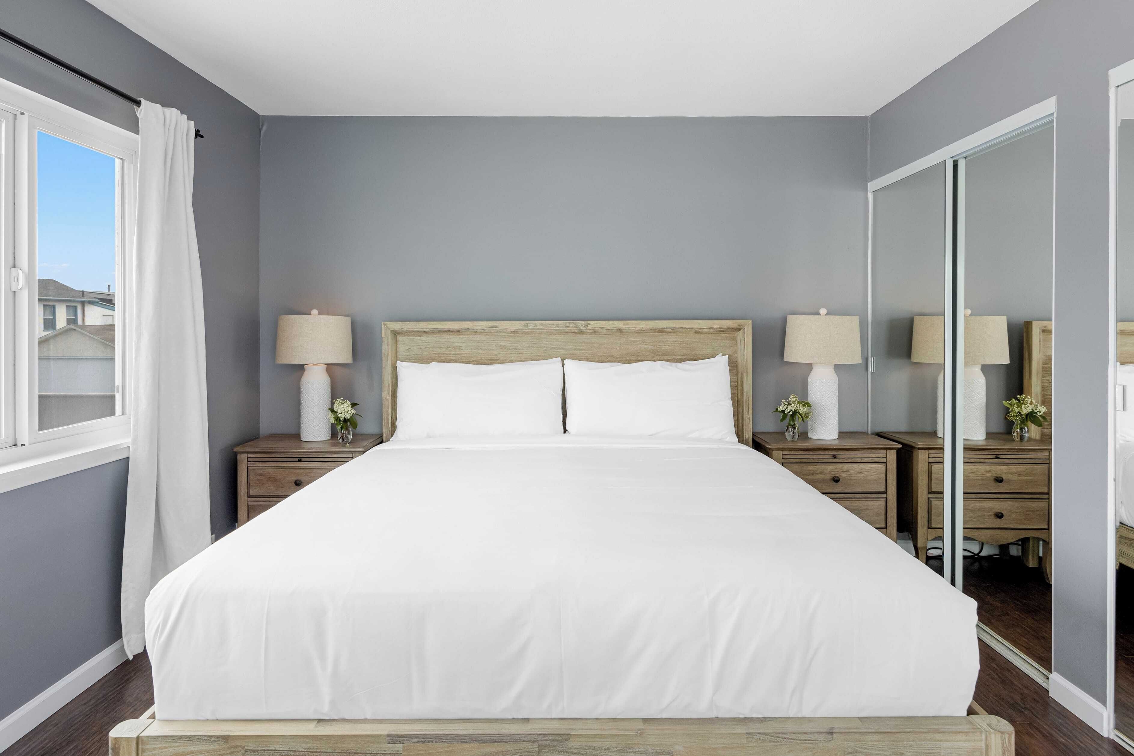 Une chambre à coucher avec un grand lit, deux tables de chevet avec des lampes, un miroir et une fenêtre avec des rideaux.