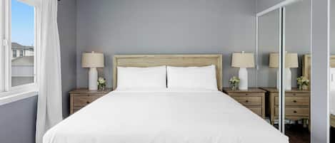 Une chambre à coucher avec un grand lit, deux tables de chevet avec des lampes, un miroir et une fenêtre avec des rideaux.