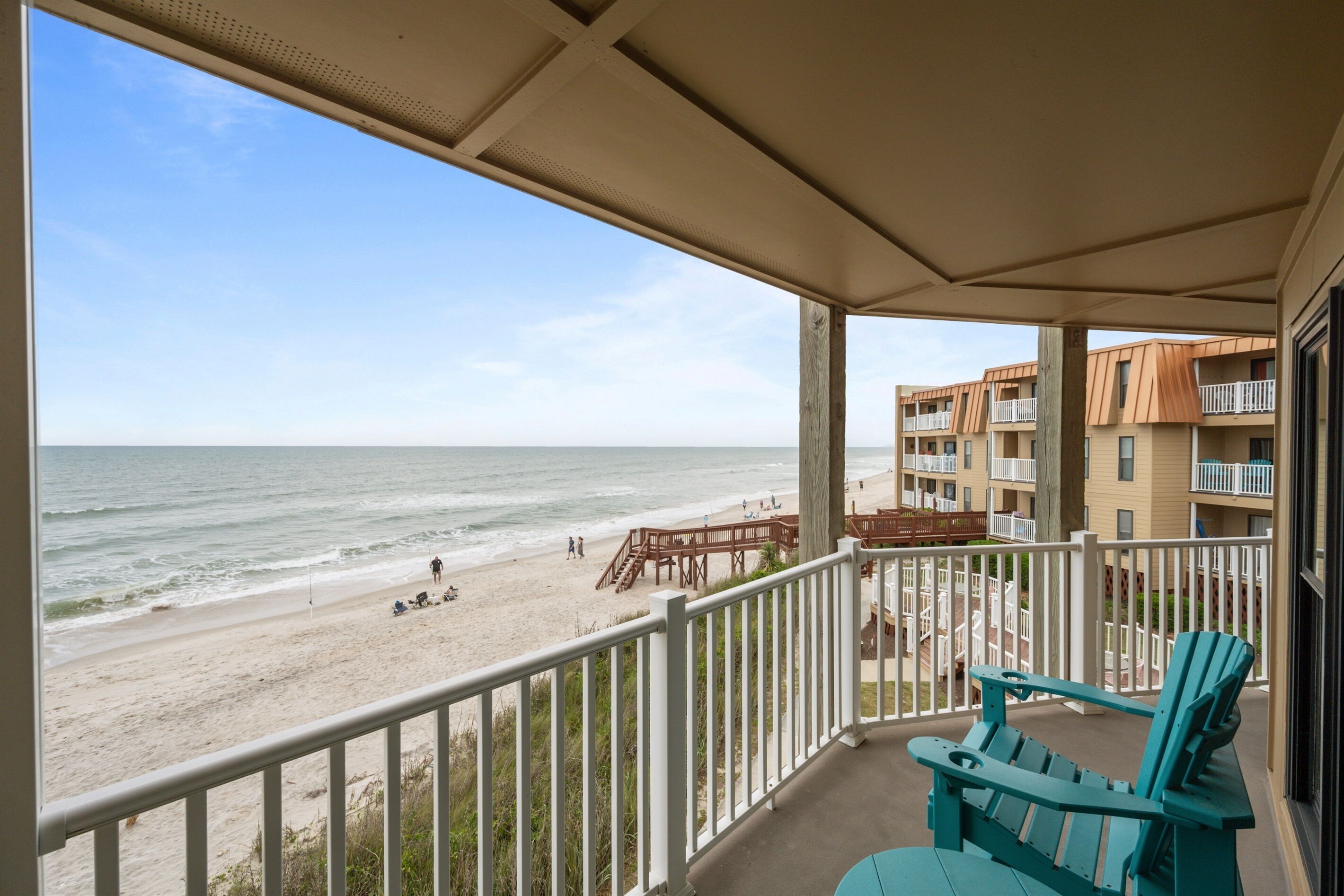A balcony with a view of the beach, wooden deck, and outdoor seating.