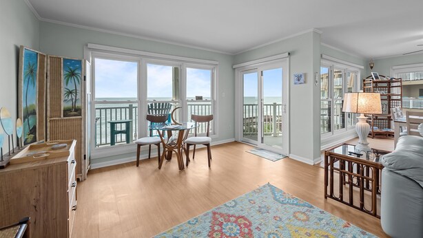 A spacious living room with a large window overlooking a balcony, wooden flooring, and a dining area with chairs and a table.