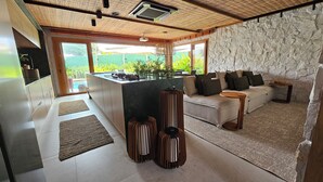A modern kitchen with a central island, a stone accent wall, and a cozy seating area with a sofa and side table. - Casa Marluá (Camacari)
