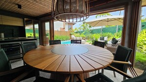 A round wooden table with chairs, a TV, and a hanging light fixture in a room with large windows and a view of a garden. - Casa Marluá (Camacari)