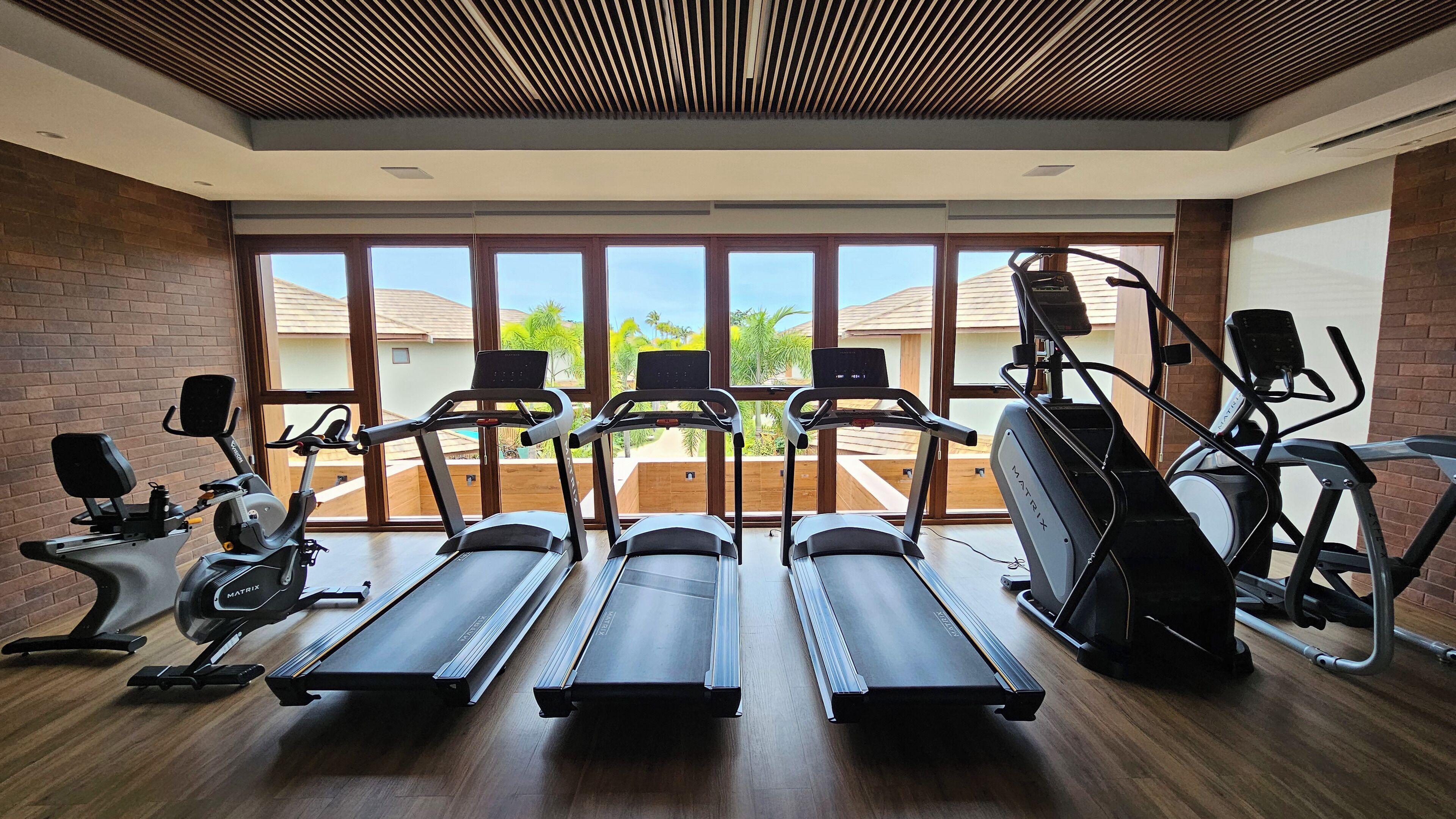 A gym with treadmills, elliptical machines, and a stationary bike, with large windows offering a view of the outdoors.