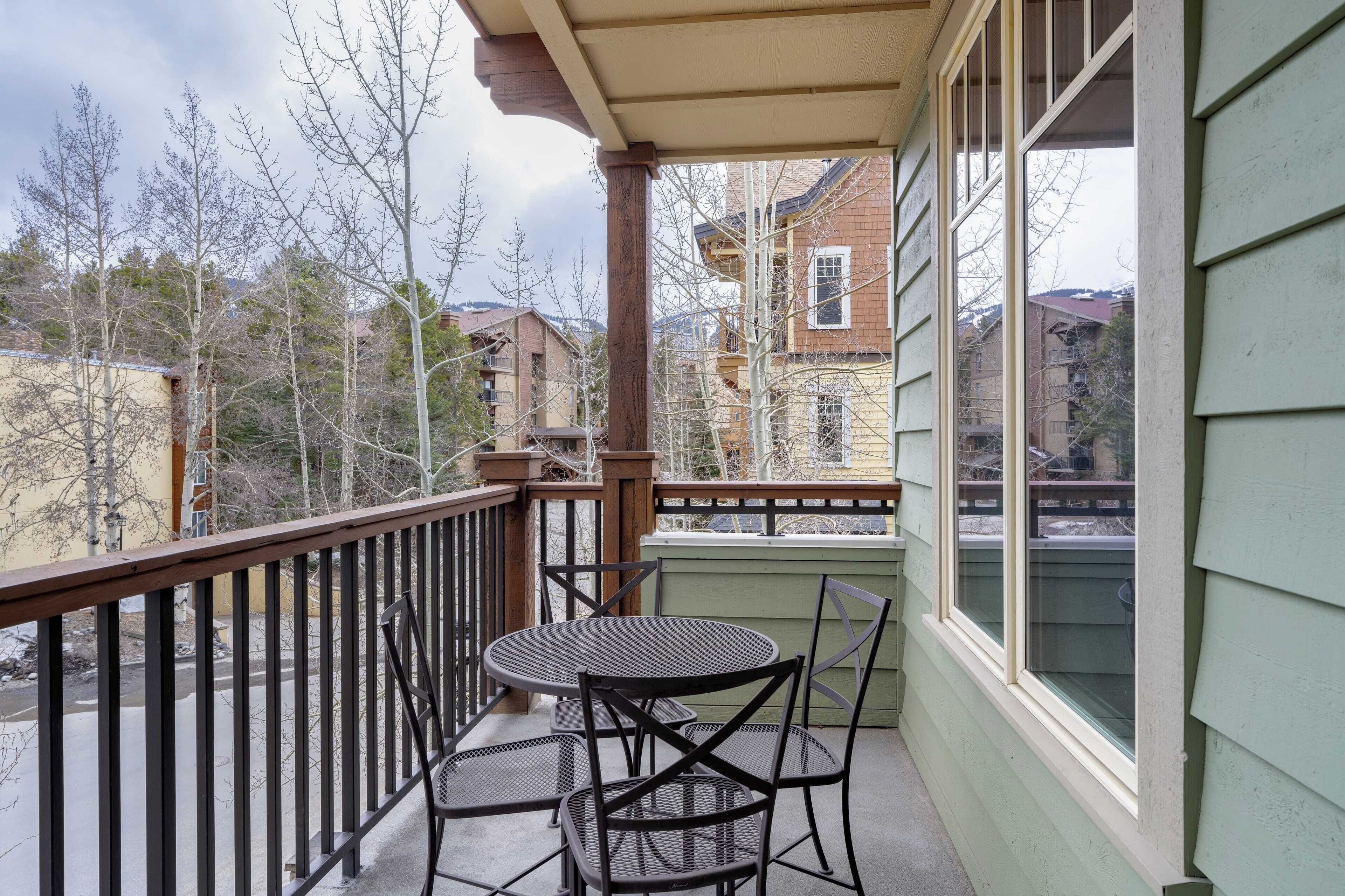 A balcony with a metal table and chairs, overlooking a residential area with trees and houses.