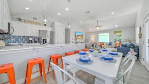 A modern kitchen with white cabinets, a central island with stools, and a dining area with blue and white plates. - Sleeps 21, 2 Homes Private Pool, Patio Lounge w/ Fire Pit, Bar, TV, & Games (Port Aransas)