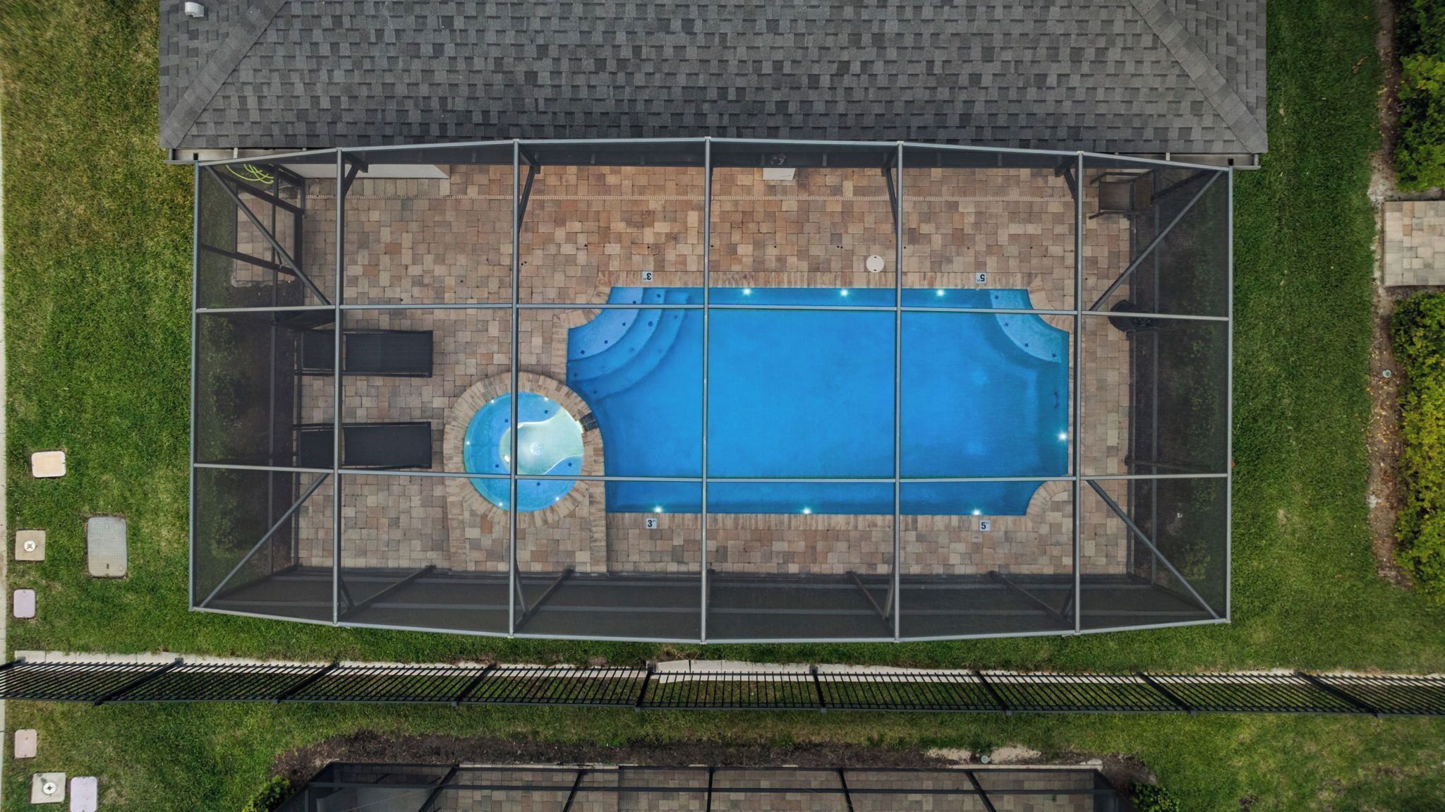 Vista aérea de una piscina con zona de cubierta, rodeada por un muro de ladrillo y una cerca de metal.