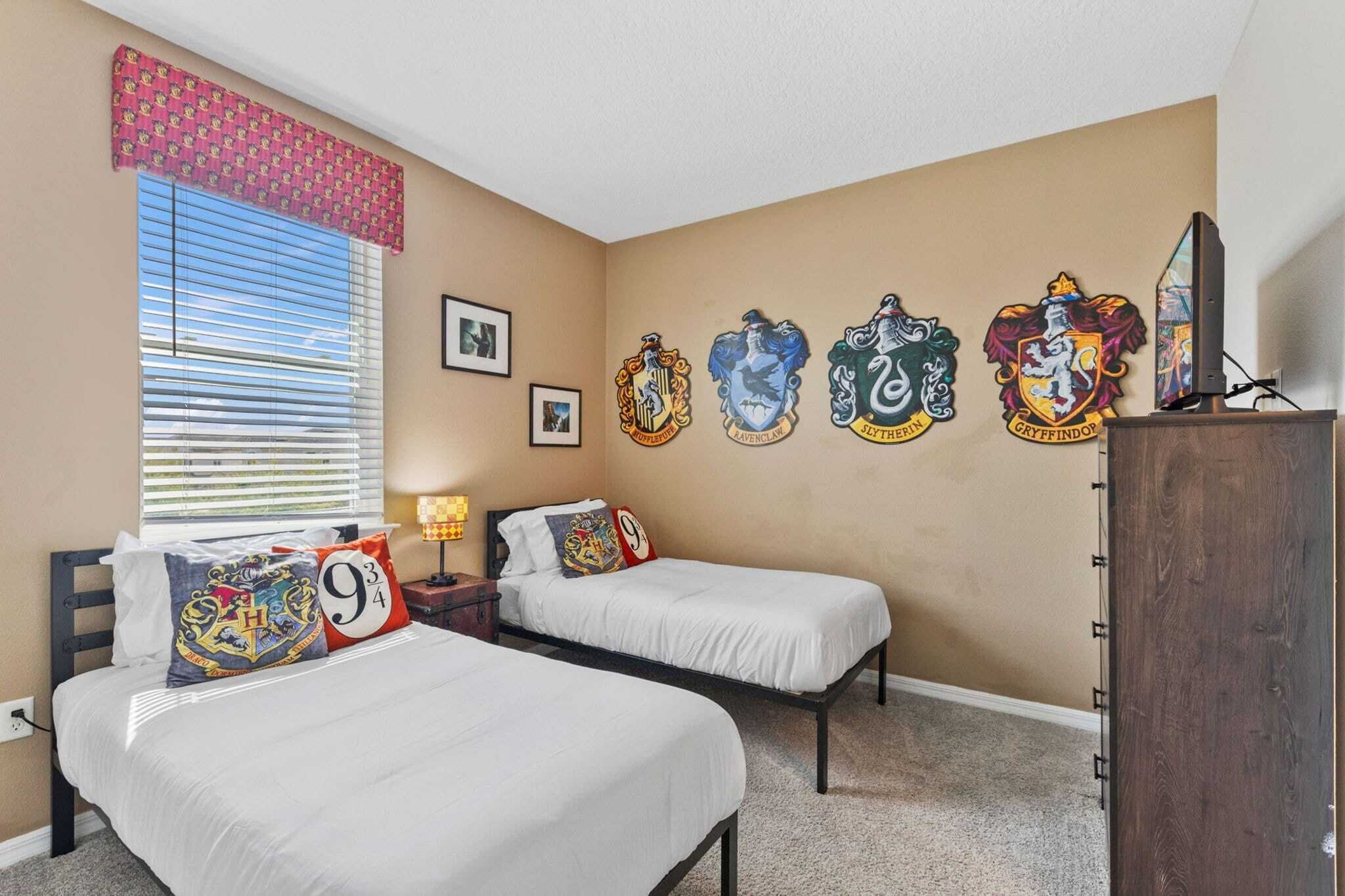 Un dormitorio con dos camas, una ventana con persianas, una pared con cinco escudos de las casas de Hogwarts y un televisor sobre un mueble de madera.