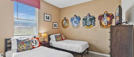 Un dormitorio con dos camas, una ventana con persianas, una pared con cinco escudos de las casas de Hogwarts y un televisor sobre un mueble de madera.