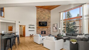 A spacious living area with a stone fireplace, a large window with a view of trees, and a modern kitchen with bar stools. - Creekside Lodge | Hot Tub | Ski-Out | Pool Table (Panorama)