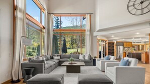 A spacious living area with a large sectional sofa, grey ottomans, and a coffee table. There are large windows with blinds, a view of greenery outside, and a kitchen area visible through an open door. - Creekside Lodge | Hot Tub | Ski-Out | Pool Table (Panorama)