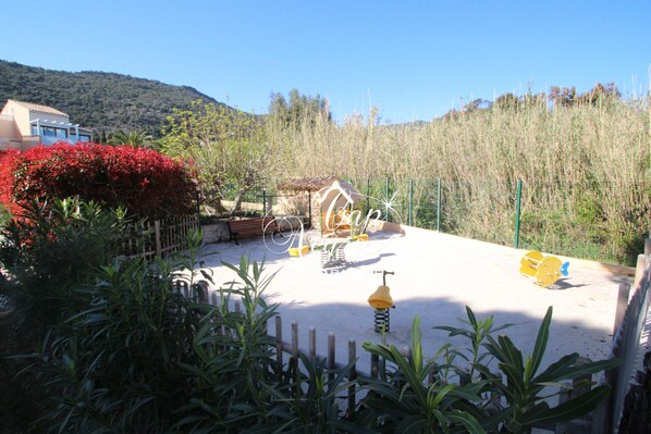 Un espacio al aire libre arenoso con una pequeña cabaña, un arenero amarillo y un arbusto rojo con flores.