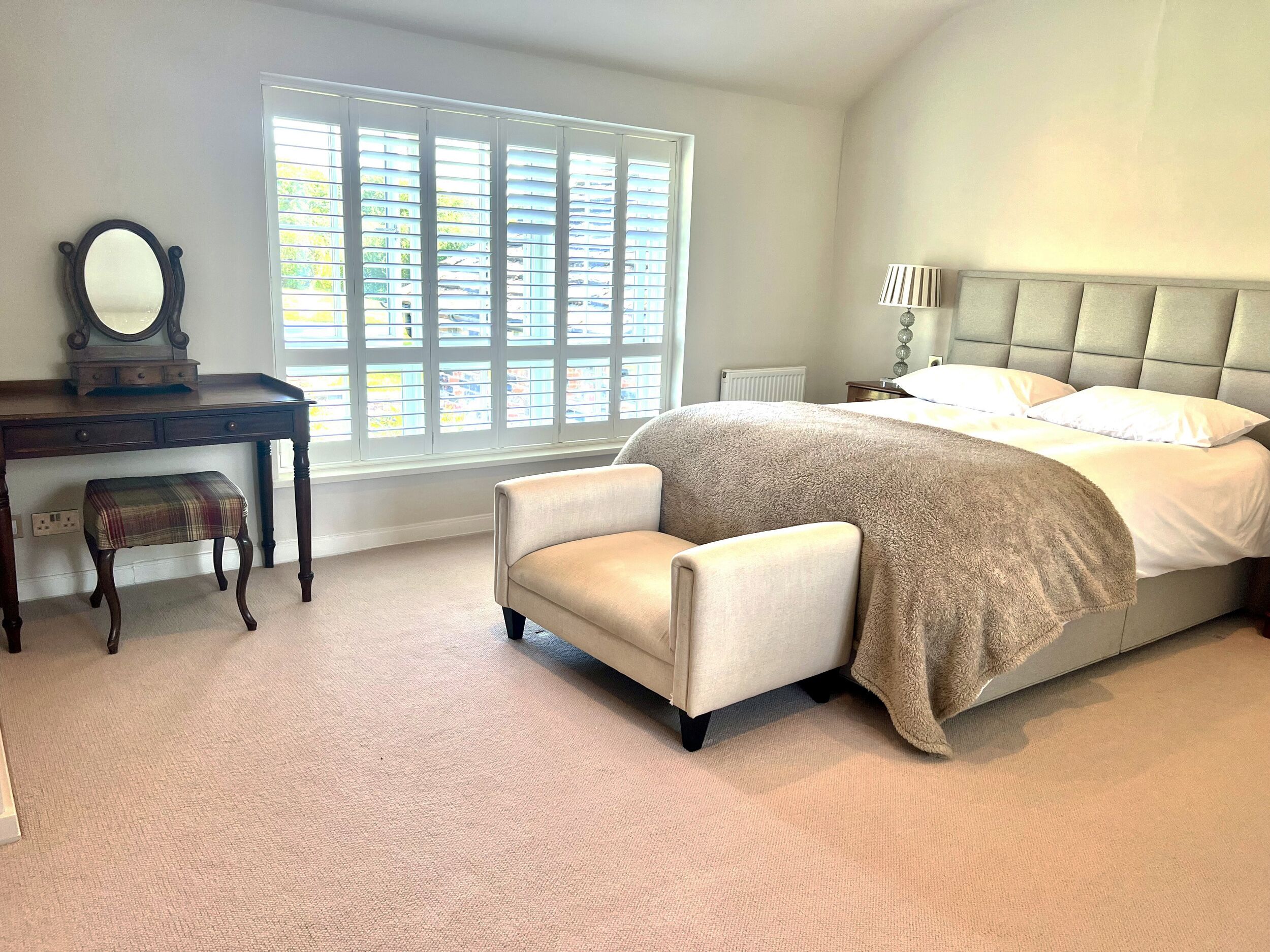 A bedroom with a bed, a desk with a mirror, a bench, and a window with blinds.