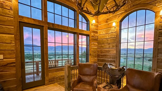 A log cabin interior with large arched windows, a patterned rug, leather chairs, and a deer antler chandelier. - Rustic Luxury Barn Retreat in Pray, Montana – Family & Pet Friendly (Livingston)