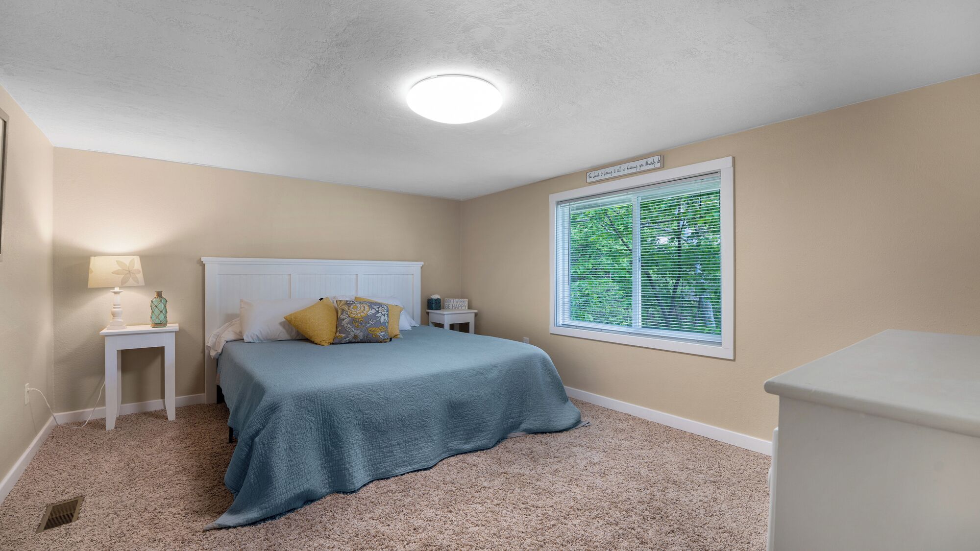 A bedroom with a bed, a nightstand, a lamp, a window with blinds, and a ceiling light.