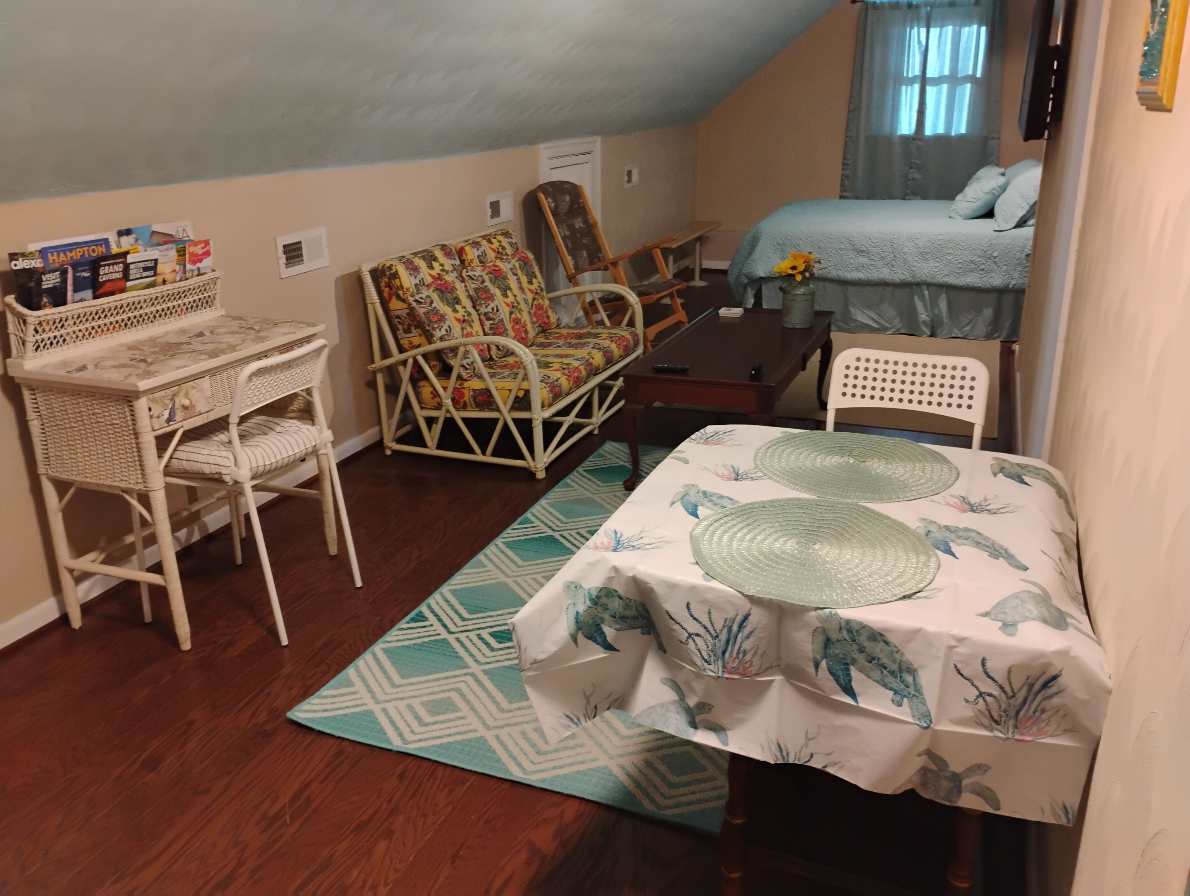 Una habitación compacta con cama, un escritorio pequeño, una silla de mimbre y una mesa de comedor.