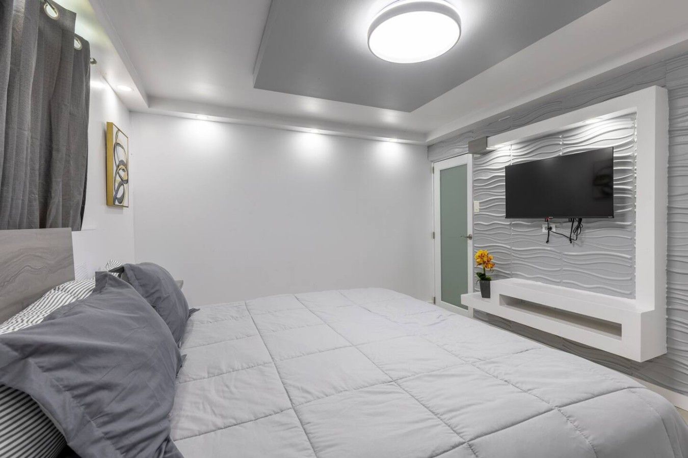 A modern bedroom with a bed, a flat-screen TV mounted on a textured wall, and a ceiling light.