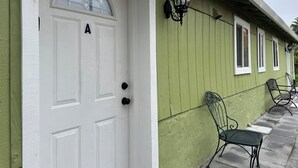 Eine weiße Tür mit Glasfenster und einem schwarzen Buchstaben 'A', eine grüne Außenwand und zwei Metallstühle auf der Veranda.
