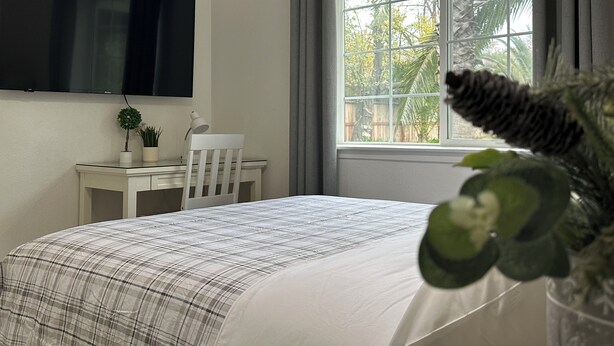 Ein Schlafzimmer mit Bett, Schreibtisch, Stuhl, Fernseher und einem Fenster mit Vorhängen.